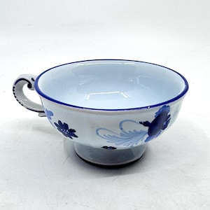 May include: White ceramic teacup with a blue floral design. The cup has a rounded bowl shape, a handle, and a pedestal base. The rim and handle are outlined in blue. The floral pattern includes stylized flowers and leaves.