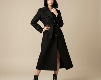 Può includere: Un trench nero lungo con cappuccio, doppiopetto, cintura e spacco alto. Il cappotto è indossato con stivaletti neri. La modella indossa il cappotto.