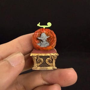 Peut inclure: Petite sculpture représentant un personnage dans une sphère orange flamboyante, posée sur un coffre décoratif. Le personnage porte un chapeau et un collier. Le coffre a des détails dorés et un coussin rouge. La sphère a une tige verte.
