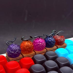 May include: A collection of colorful, stylized fruit-shaped figurines. Each figurine is a different color, including purple, orange, pink, blue, and red. They have a textured surface and are mounted on small, brown platforms.