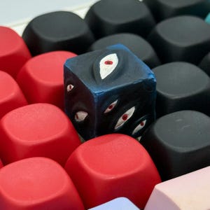 May include: A custom artisan keycap featuring a cube design with multiple painted white eyes with red pupils. The cube is surrounded by red, black, and light pink keycaps, creating a unique and artistic keyboard accessory.