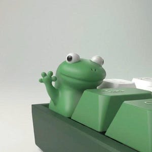 May include: A green frog ornament emerging from a green keyboard. The frog has large, white eyes and is waving a small hand. The keyboard keys are green, with white keys visible in the background.