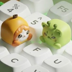 May include: Two custom mechanical keyboard keycaps shaped like small characters. One is a yellow, pumpkin-like figure with a cat face, and the other is a light green, rounded creature with antennae, both resting on white keyboard keys with black letters.