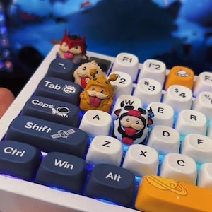 Könnte beinhalten: Eine mechanische Tastatur mit weißen und dunkelblauen Tastenkappen, die mit Text und Symbolen bedruckt sind. Mehrere individuelle Artisan-Tastenkappen in Form kleiner Zeichentrickfiguren, darunter eine Kuh und eine Biene, sind zwischen den Tasten.