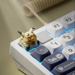 May include: A custom mechanical keyboard keycap featuring a small yellow character wearing a beige hooded costume with black ear tips. The keycap is installed on a white keyboard with blue backlighting, positioned near the Tab and F1 keys.