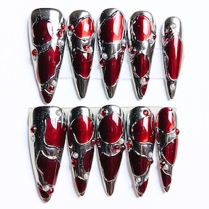 May include: Set of ten long, pointed false nails. Each nail features a silver metallic frame with a deep red, glossy center. Embellishments include small red and white gems. The nails are arranged on a clear plastic holder.