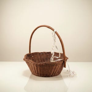 May include: A round, dark brown woven wicker basket featuring a single arched handle. A thin, white, curled decorative ribbon is tied to the handle and hangs down into the interior and over the side of the basket, which has a textured weave.