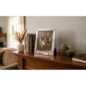May include: A rectangular silver-toned picture frame with decorative corner accents, containing a black and white vintage photograph of a family group. The frame is positioned upright on a wooden surface next to books and a glass terrarium.