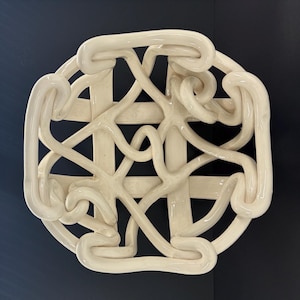 Puede incluir: Una escultura mural circular de color crema con un diseño complejo e interconectado. La escultura presenta una base similar a una rejilla superpuesta con formas tubulares en bucle, creando un efecto texturizado y tridimensional. La pieza está montada sobre un fondo azul oscuro.