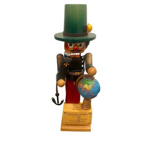 May include: A wooden nutcracker figure wearing a tall green hat, holding a small black bird and a globe. The base features a plaque with the text: Christoph Columbus, America 1492, 500 years ago, Memorial 1992, Times, Fortune and Faith with him.