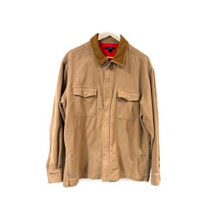 May include: A tan long-sleeve button-up shirt featuring a brown corduroy collar and a bright red inner lining. The shirt includes two chest pockets with buttoned flaps and a J.Crew brand label visible on the inside of the collar area.
