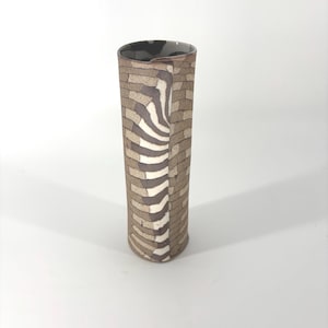 May include: A tall, cylindrical ceramic vase with a unique design. The vase features a pattern of rectangular shapes in shades of brown and tan, with a vertical zebra-like stripe. The interior rim is dark with a mottled pattern.