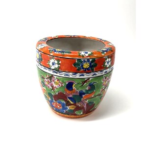 May include: A ceramic planter featuring a vibrant green base decorated with colourful birds and floral patterns. The upper rim is a wide orange band adorned with small white and blue flowers. The interior of the vessel is a smooth, neutral grey.