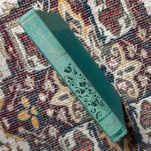 May include: A green hardcover book with the title "The Little Prince" on the spine. The book has a gold-colored design on the spine and a green and black floral design on the front cover. The book is by Antoine de Saint-Exupéry and published by Knopf.