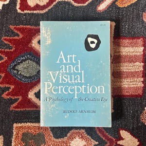 May include: A book titled "Art and Visual Perception" by Rudolf Arnheim. The cover is light blue with white text and a black and white abstract design. The book is on a patterned rug.