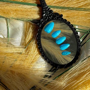 May include: A black, ornate hand mirror reflects five turquoise-coloured, almond-shaped artificial nails. The nails are arranged in a row, with the smallest nail at the bottom. The mirror is set against a backdrop of yellow, feathery material.