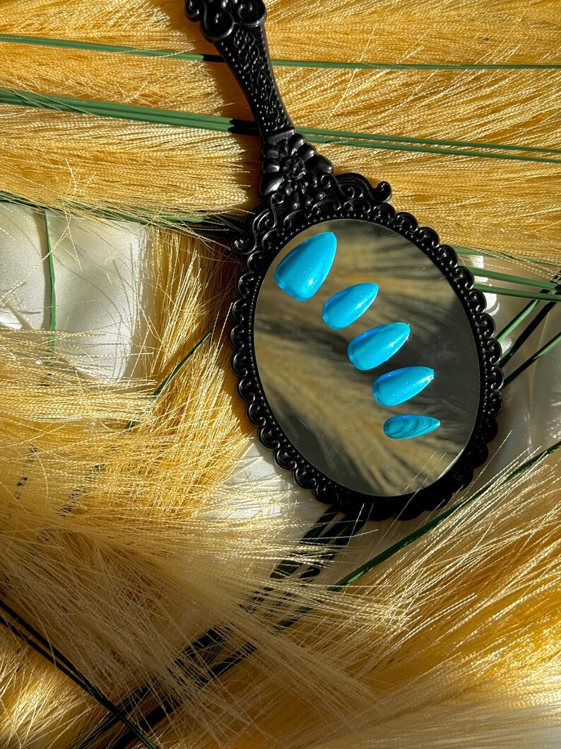 May include: A black, ornate hand mirror reflects five turquoise-coloured, almond-shaped artificial nails. The nails are arranged in a row, with the smallest nail at the bottom. The mirror is set against a backdrop of yellow, feathery material.