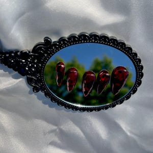 May include: An oval black hand mirror reflects five long, glossy, dark red artificial nails. The nails have a marbled appearance. The mirror has a decorative, ornate frame. The background is a white, silky fabric.