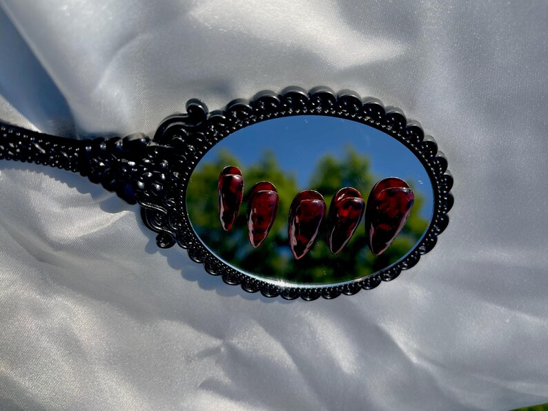 May include: An oval black hand mirror reflects five long, glossy, dark red artificial nails. The nails have a marbled appearance. The mirror has a decorative, ornate frame. The background is a white, silky fabric.