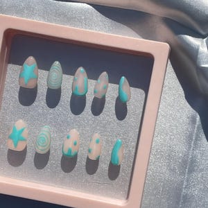 May include: A set of ten artificial nails in a pink case. Each nail is a light beige color with a different design in turquoise. Designs include a star, swirls, dots, and curved lines. The nails are oval-shaped.