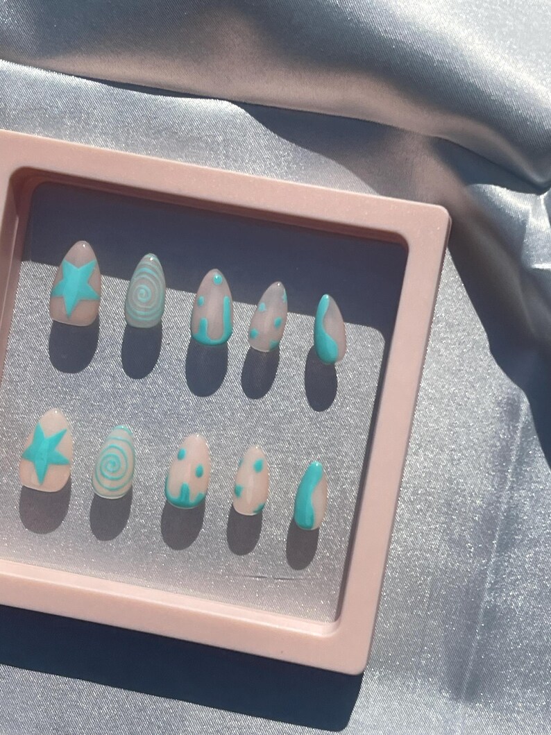 May include: A set of ten artificial nails in a pink case. Each nail is a light beige color with a different design in turquoise. Designs include a star, swirls, dots, and curved lines. The nails are oval-shaped.