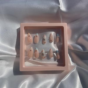 May include: A collection of artificial nails presented in a pink display case. The nails are a natural pink hue, some adorned with tiny silver rhinestones, others with light blue tips or small star motifs. The display case is a pale pink.