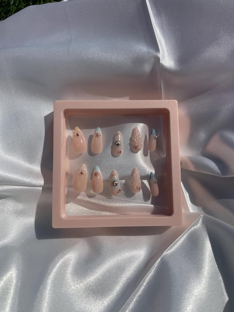 May include: A collection of artificial nails presented in a pink display case. The nails are a natural pink hue, some adorned with tiny silver rhinestones, others with light blue tips or small star motifs. The display case is a pale pink.