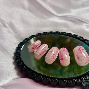 May include: Oval-shaped, pink and white press-on nails with a decorative design. The nails are arranged on a black, ornate mirror, set against a pink satin background. The design features swirls and lines.