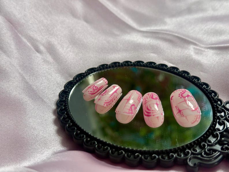 May include: Oval-shaped, pink and white press-on nails with a decorative design. The nails are arranged on a black, ornate mirror, set against a pink satin background. The design features swirls and lines.
