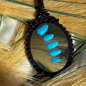 May include: A black, ornate hand mirror reflects five bright blue, oval-shaped artificial nails. The mirror has a decorative, scalloped edge. The background features dried, yellow-toned grasses.