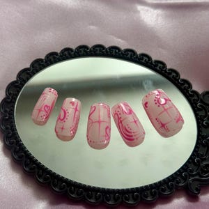 May include: Set of six pink press-on nails with a glossy finish. Each nail features a pink design with hearts, lines, and dots. The nails are displayed on a black framed mirror.