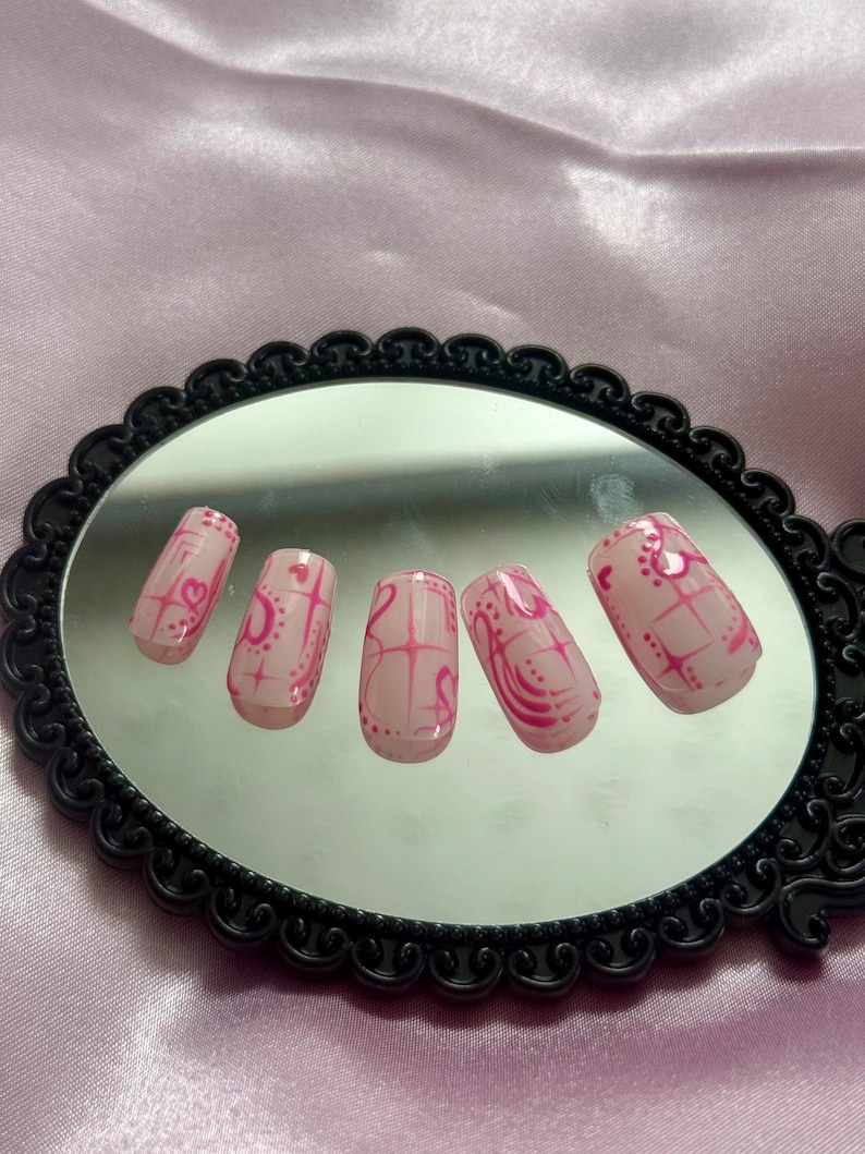 May include: Set of six pink press-on nails with a glossy finish. Each nail features a pink design with hearts, lines, and dots. The nails are displayed on a black framed mirror.