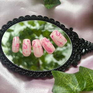 May include: A set of five artificial nails, each painted with a light pink base and a vibrant pink design. The design includes hearts, lines, and dots. The nails are displayed on a black, ornate mirror.