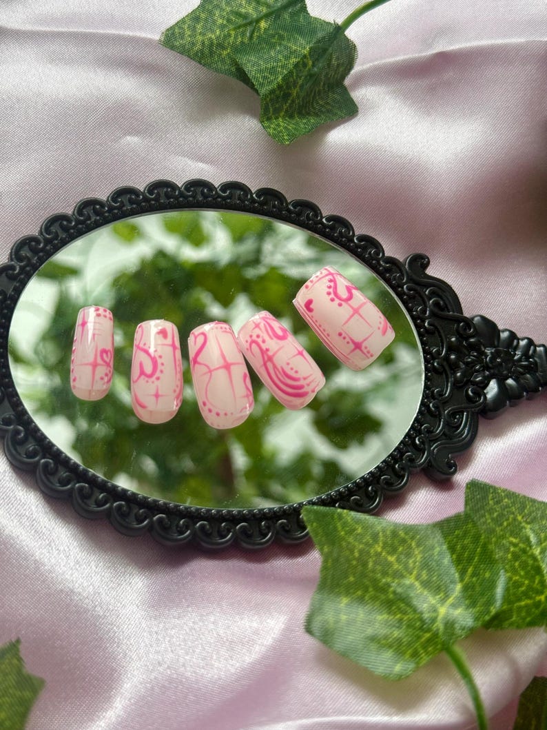 May include: A set of five artificial nails, each painted with a light pink base and a vibrant pink design. The design includes hearts, lines, and dots. The nails are displayed on a black, ornate mirror.