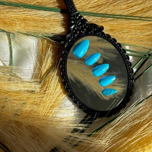 May include: A black, ornate hand mirror reflects five bright blue, almond-shaped artificial nails. The mirror is set against a backdrop of yellow-toned, feathery decorative elements, creating a contrasting visual texture.