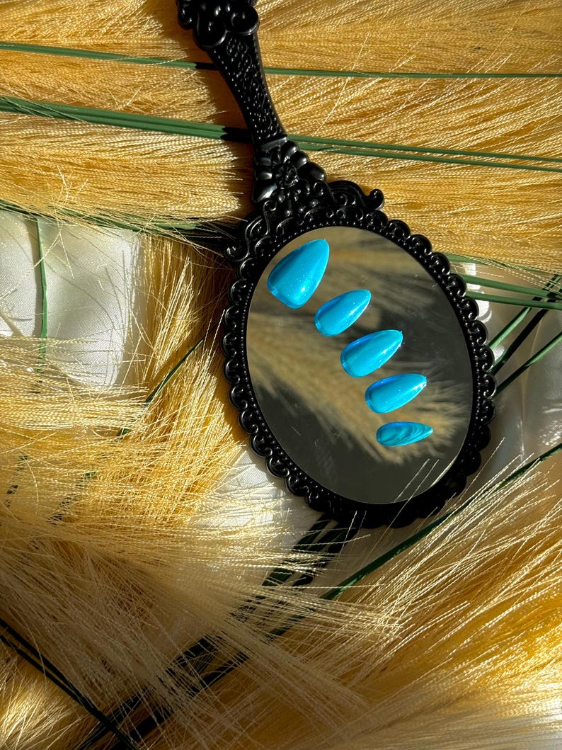 May include: A black, ornate hand mirror reflects five bright blue, almond-shaped artificial nails. The mirror is set against a backdrop of yellow-toned, feathery decorative elements, creating a contrasting visual texture.