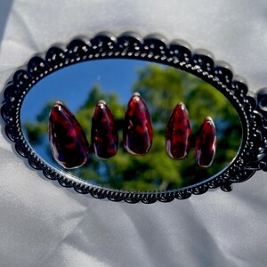 May include: Five almond-shaped artificial nails with a dark red and brown tortoiseshell pattern are displayed on a decorative black mirror. The mirror reflects a blurred outdoor scene. The nails are arranged in a row.