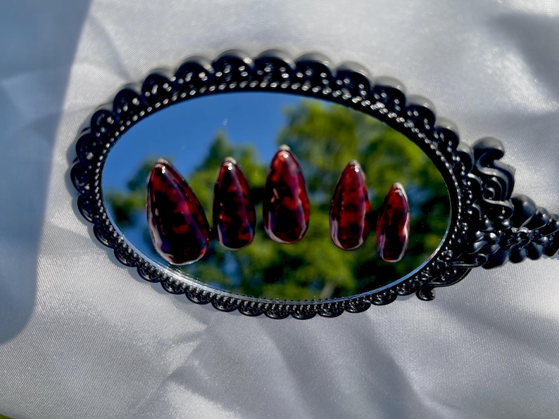 May include: Five almond-shaped artificial nails with a dark red and brown tortoiseshell pattern are displayed on a decorative black mirror. The mirror reflects a blurred outdoor scene. The nails are arranged in a row.