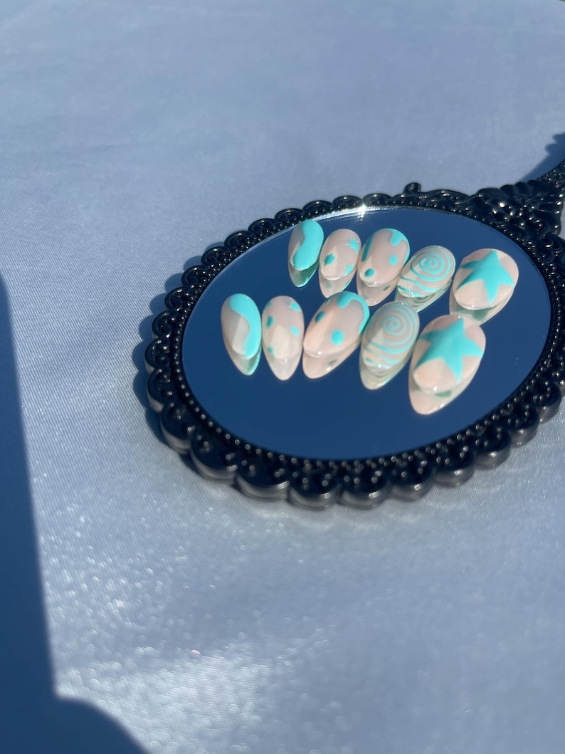 May include: A set of ten almond-shaped false nails. Each nail is painted with a nude base and adorned with light blue designs, including swirls, stars, and abstract shapes. The nails are arranged on a black, ornate mirror.