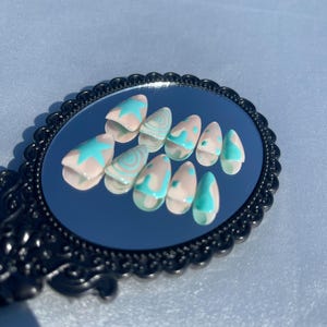 May include: A set of almond-shaped false nails displayed on a small, ornate mirror. The nails are painted with a light pink base and feature turquoise star and swirl designs. The mirror has a black, decorative frame.