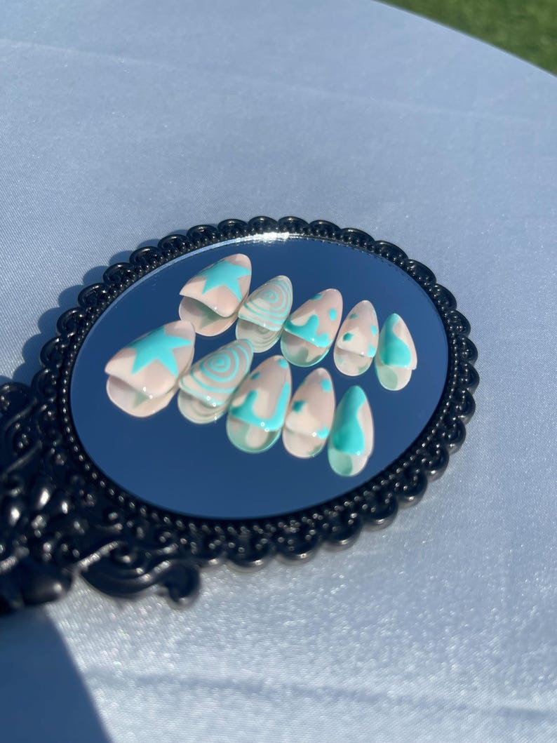 May include: A set of almond-shaped false nails displayed on a small, ornate mirror. The nails are painted with a light pink base and feature turquoise star and swirl designs. The mirror has a black, decorative frame.