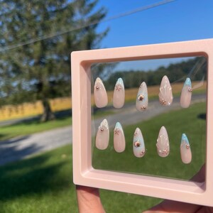 May include: A set of artificial nails in a pink display case. The almond-shaped nails have a nude base, gold embellishments, and light blue tips. The background shows a sunny outdoor scene.