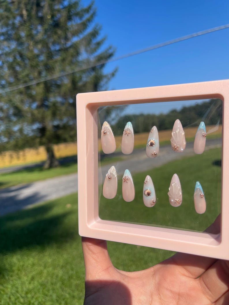 May include: A set of artificial nails in a pink display case. The almond-shaped nails have a nude base, gold embellishments, and light blue tips. The background shows a sunny outdoor scene.