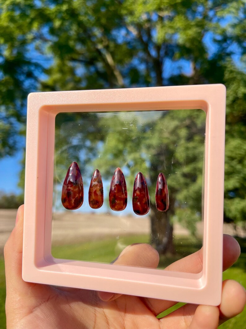 May include: A set of five artificial nails with a glossy, dark red and brown tortoiseshell design. The nails are displayed inside a light pink square frame, held by a hand. The background is a blurred view of green trees and a blue sky.