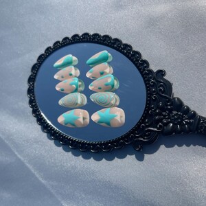 May include: A set of ten almond-shaped false nails. Each nail features a light pink base with a teal design. Designs include a star, swirls, and curved lines. The nails are displayed on a black, ornate mirror.