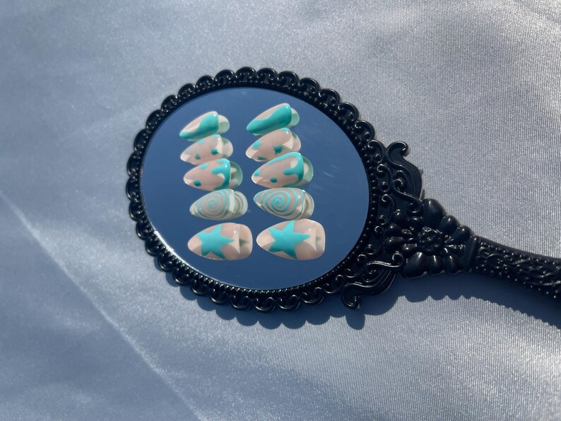 May include: A set of ten almond-shaped false nails. Each nail features a light pink base with a teal design. Designs include a star, swirls, and curved lines. The nails are displayed on a black, ornate mirror.