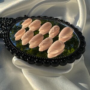 May include: A set of ten light pink, sculpted artificial nails arranged on a black-framed oval mirror. The nails have a glossy finish and a subtle, textured design. The mirror rests on a white, silky fabric.