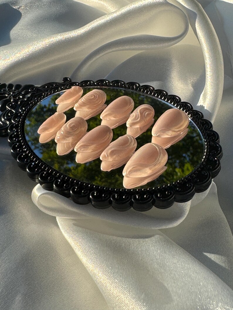 May include: A set of ten light pink, sculpted artificial nails arranged on a black-framed oval mirror. The nails have a glossy finish and a subtle, textured design. The mirror rests on a white, silky fabric.