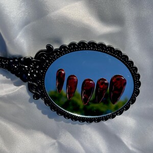 May include: An oval black hand mirror reflects five long, burgundy-colored artificial nails. The nails have a glossy finish and a marbled appearance. The mirror's frame has an ornate, scalloped design.