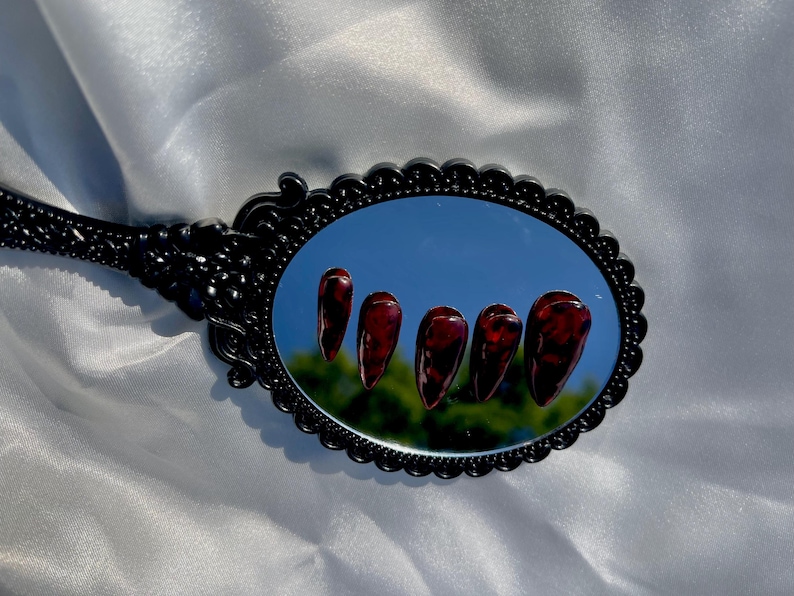 May include: An oval black hand mirror reflects five long, burgundy-colored artificial nails. The nails have a glossy finish and a marbled appearance. The mirror's frame has an ornate, scalloped design.